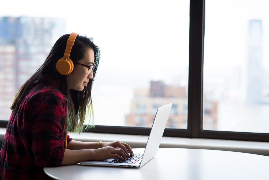 Femme-presenting Asian person with yellow headphones, red plaid shirt, sitting in front of their laptop looking at the screen.