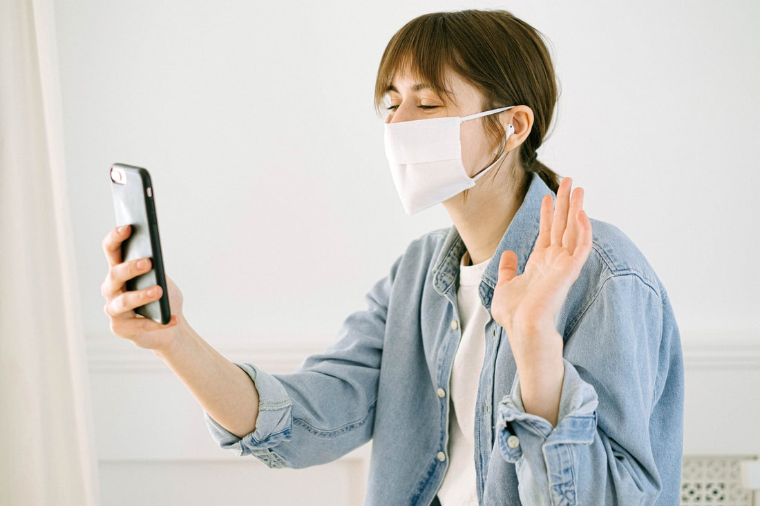 A white, brunette, femme-presenting person with white face mask wearing a jean jacket. They are waving to someone over the phone during a video call.