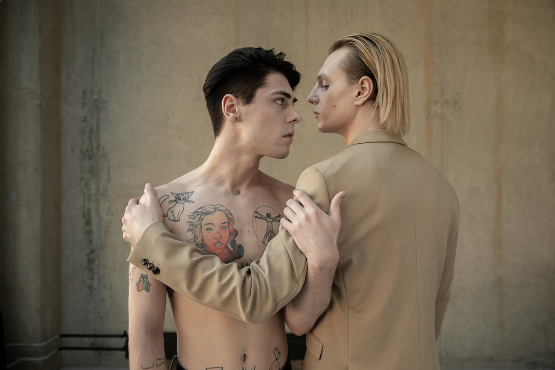 Two white, masculine-presenting people embracing one another by the arm. The person on the left has several tattoos across the chest, including a cartoon cat, a colorized smiling woman, and da Vinci's Vitruvian Man. The person on the left is wearing a tan blazer.