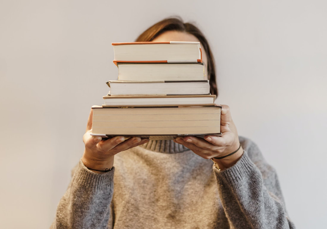 Femme-presenting person with a stack of books in their hands. They are held over their face. The person is wearing a gray sweater in warm light in front of a light grey background.