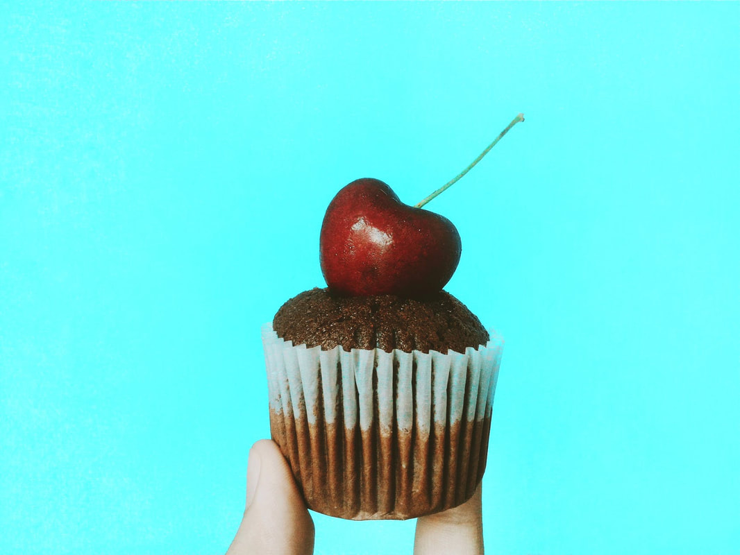 Small chocolate cupcake held up against a turquoise background. There is a cherry on top of the cupcake.