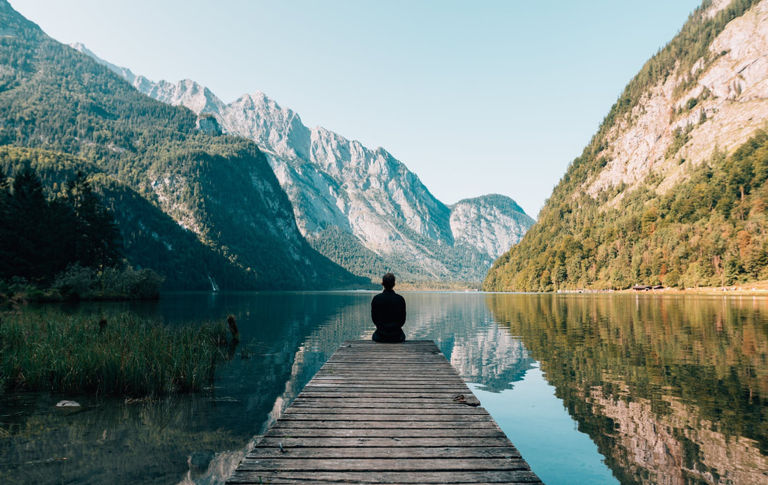 Person sitting at the end of a dock next to forests and mountains. 